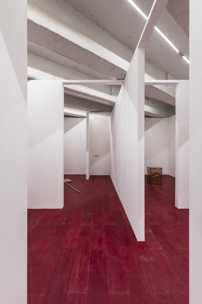 immagine per Pavimento, 2023 Cera, legno, cartongesso; cm 600x700x3 Una struttura in cartongesso, di metri 6 per 7, è la riproduzione in scala 1:1 della pianta della hutte che Martin Heidegger si fece costruire nei pressi di Todtnauberg nella Foresta Nera in Germania. In questa capanna Heidegger era solito ritirarsi con sua moglie per studiare e scrivere a contatto con la natura del luogo, la hutte è famosa anche perché è stato luogo di incontro tra Heiddeger e numerose personalità dell’epoca, famoso resterà l’incontro del 26 luglio 1967 tra il filosofo e il poeta Paul Celan. L’opera riproduce solo le dimensioni e la sistemazione degli spazi interni della hutte originale, non sono riportate le finestre e neanche il tetto della capanna, l’altezza delle pareti della scultura corrisponde all’altezza tra il pavimento e il soffitto dello spazio che accoglie l’opera. Per accedere all’interno della scultura c’è un gradino alto 15 centimetri, l’esterno della scultura e le pareti interne sono dipinte di un colore quanto più simile possibile a quelli delle pareti dello spazio, il pavimento della scultura è composto da una riproduzione in cera da fonderia rossa di un asse di legno lunga 100 centimetri e larga 20, l’asse è sempre la stessa ripetuta identica per 210 volte, tanto quanto occorre per ricoprire il pavimento interno della scultura.